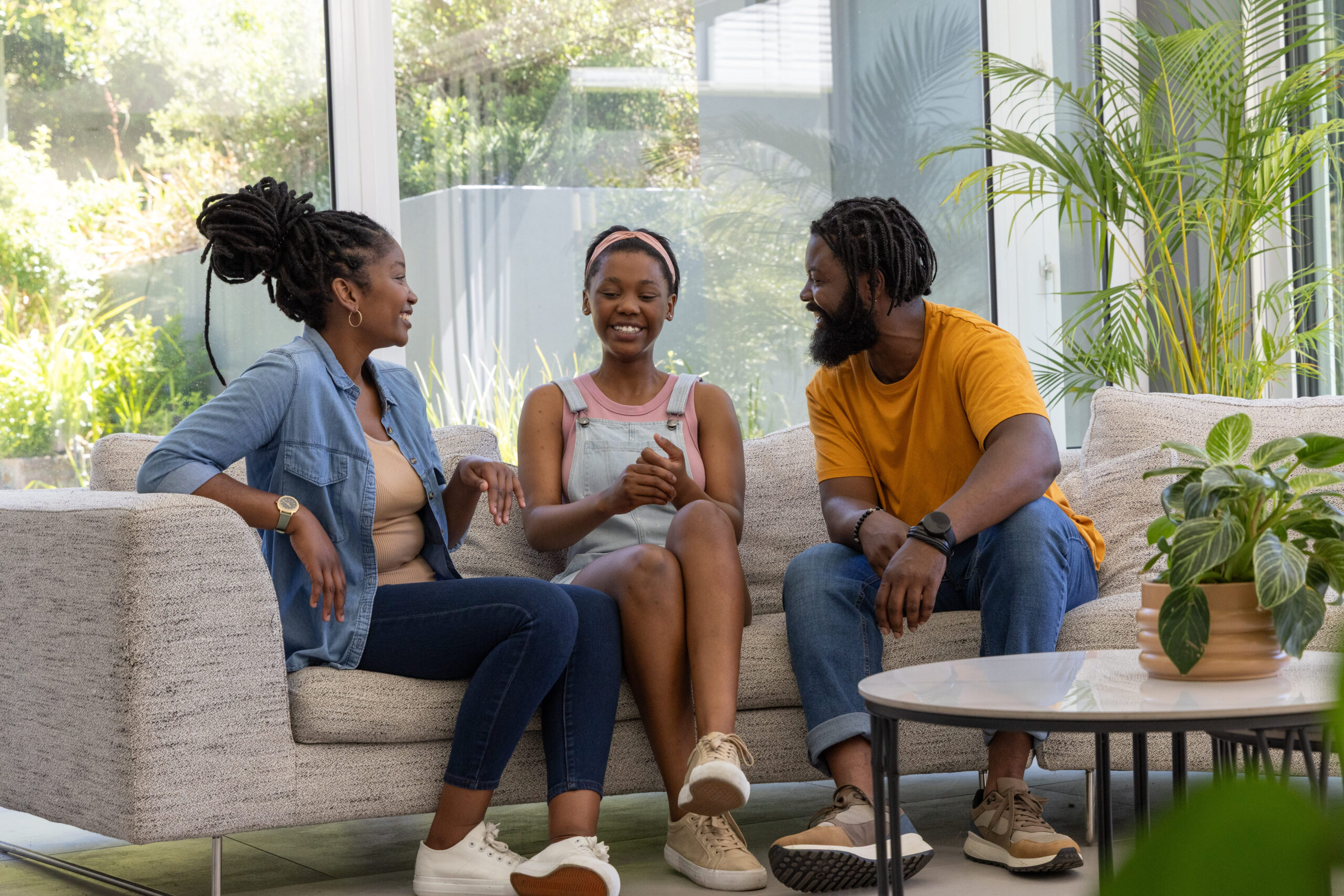 African American family sitting on light-gray sofa in modern living room with potted plants. Couch, sunlit, greenery, garden, group, conversation, home