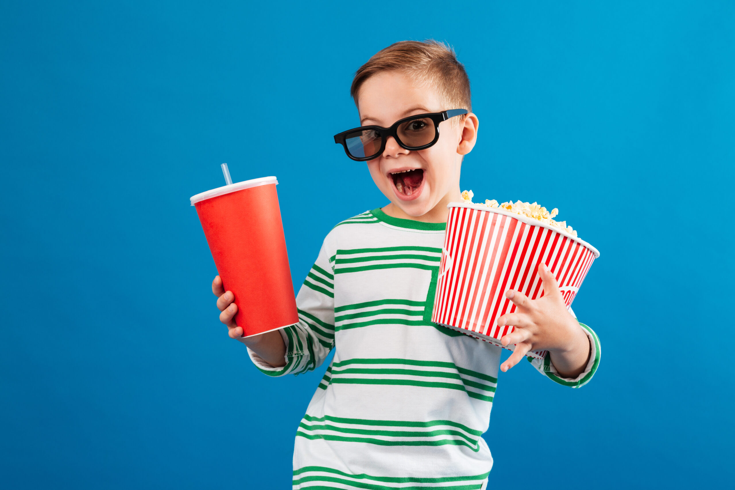 Cheerful young boy in eyeglasses preparing to watch the film while holding soda and popcorn over blue background